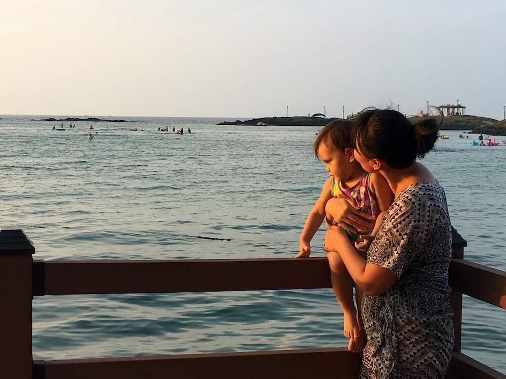 A mother holding her young daughter by the ocean, reflecting on her childhood growth before visiting a growth clinic in Seoul.