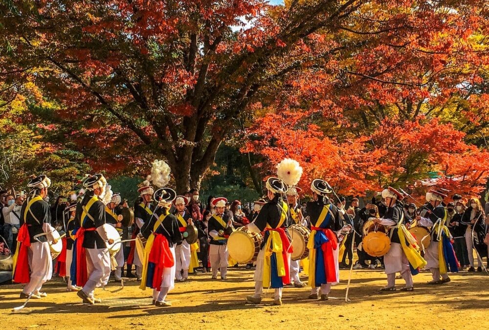 Traditional Korean musicians performing in colourful yellow and blue attire under a canopy of deep red autumn leaves at Nami Island.