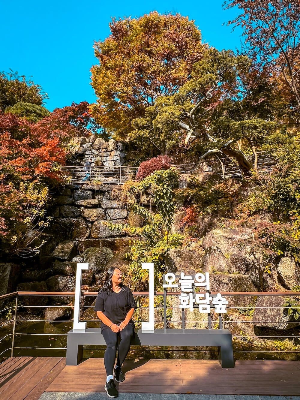 Woman sitting on a modern black photo bench in front of a stone wall waterfall and autumn trees at Hwadam Botanic Garden
