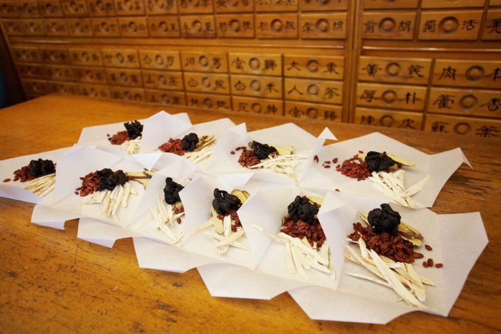 Small bundles of raw traditional Korean medicinal roots, barks, and dried herbs arranged neatly on folded sheets of white paper.