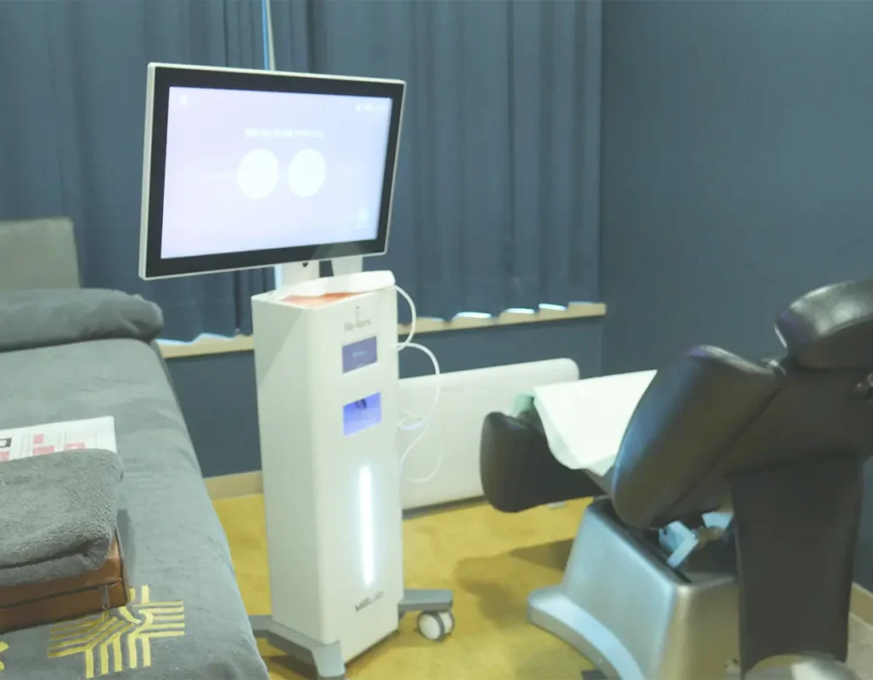 A sleek, white electronic biometric tracking machine with an overhead display screen stands next to a treatment chair.